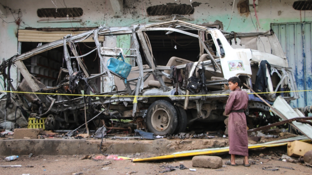 A child stands near the mangled wreckage of the bombed-out school bus [Ahmad Algohbary/Al Jazeera]