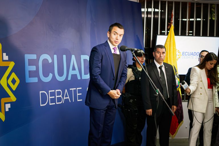 A man, Daniel Noboa, leans forward to speak into a microphone placed upon a debate stage. Behind him, a banner reads: Ecuador debate.