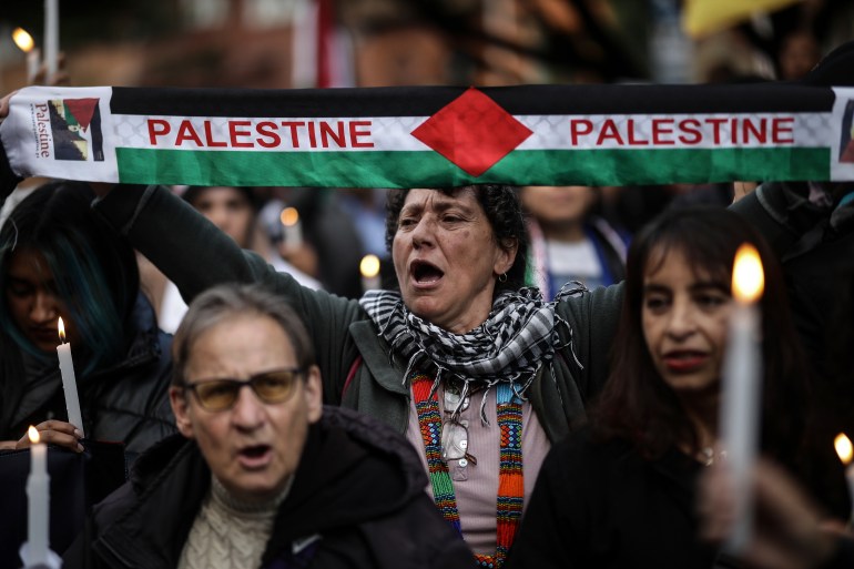 A woman holds up a scarf in the colors of the Palestinian flag, with the word "Palestine" written in red. Around her, people carry candles.