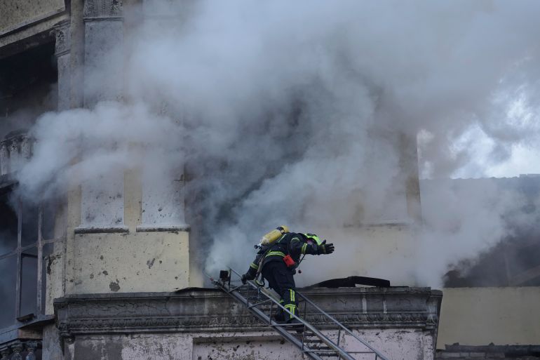 A firefighter on a ladder in front of a Kyiv building hit by a Russian missile. There is thick smoke. The firefighter is signalling to someone on the ground.