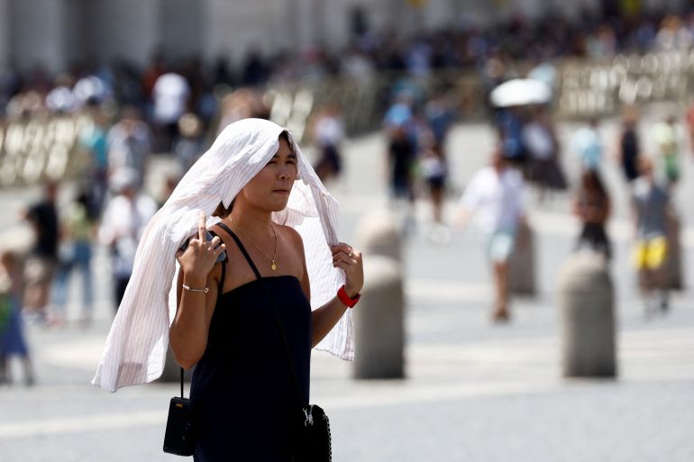 A woman covering her head with a white shirt as she walks in a square beneath the sun during a heatwave
