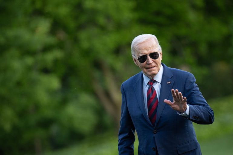 Joe Biden walking across the lawn at the White House. He is wearing a suit and aviator sunglasses. He is waving