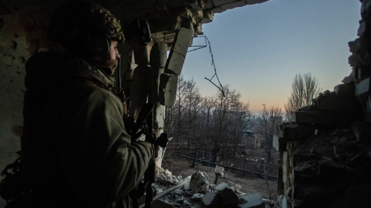 Ukrainian soldier stands in a cave, looking out towards the opening that shows a clear dimming blue sky, with a setting sun over naked trees on the horizon. It looks cold
