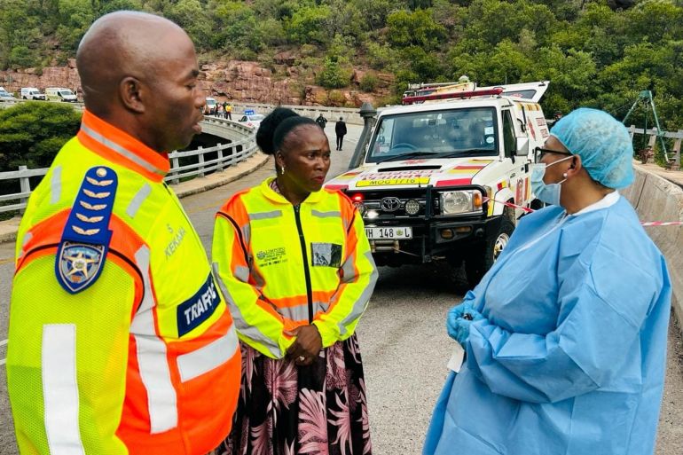 Emergency teams on the bridge where the bus crashed. Two are wearing high-viz uniforms. One is wearing pale blue medical overalls, a hat and gloves. There is a truck behind them.
