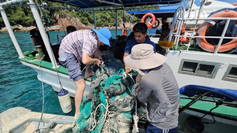 Um grupo de pessoas transportando um enorme emaranhado de redes de pesca descartadas para um pequeno barco.  As redes são verdes e cinzentas e têm nós. 