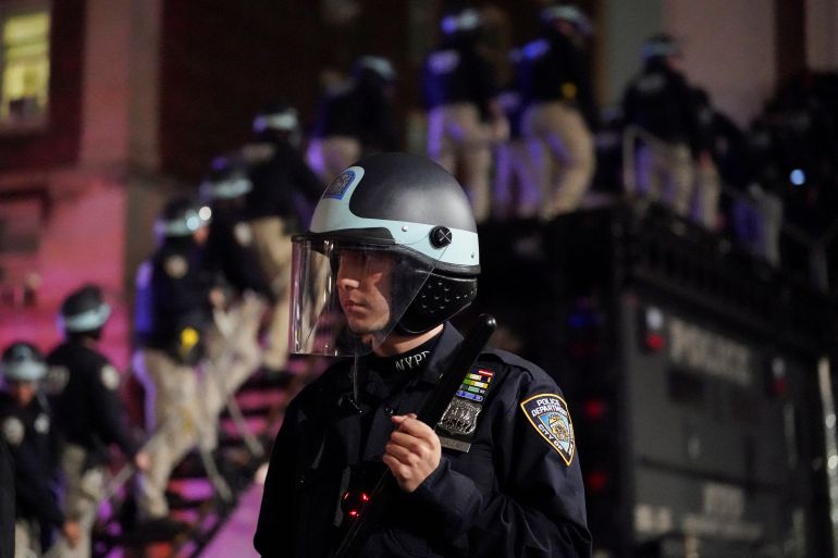 A New York police officer in a helmet. Behind him a line of police are walking up a ladder attached to a truck.